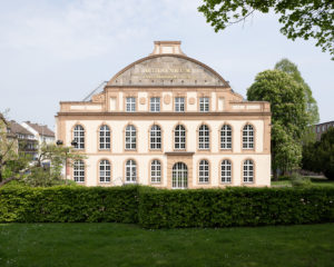The photo shows the facade of the Ottoneum. The multi-storey building consists of many windows with round arches. In front of the building is a small garden.