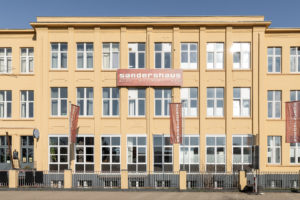 A sand-colored building with a red banner on the Sandershaus stands.