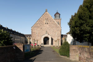 A brick church in the middle of the residential area. Tree trunks on the right and left decorate the path to the entrance.