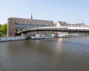 Eine Fußgänger*innenbrücke mit Sitzgelegenheiten führt über den Fluss Fulda zu einem alten Gebäude aus Stein. Am Ufer liegen ein paar Schiffe. 