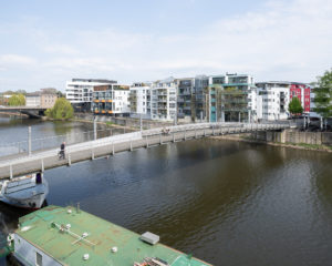 Eine Fußgänger*innenbrücke mit Sitzgelegenheiten führt über den Fluss Fulda zu einem modernen Wohngebiet. 