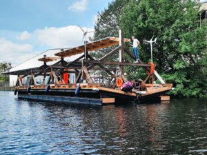 The wooden ship on the river, a man standing on a beam. In the background green, blue sky light clouds.