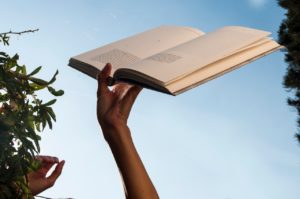 An open book is held by two hands in the blue sky. Green leaves on the left and right edge suggest trees.