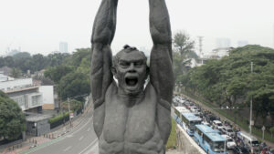 Eine Skulptur, die einen schreienden Mann mit ausgestreckten Armen darstellt. Im Hintergrund eine belebte Straße.