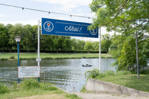 The photo shows the bank of the Fulda with a view to the river on a sunny day. Above a small concrete pier is a large blue sign with the words 'Ahoi!' supported by two poles.