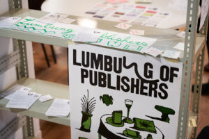Ein Stahlregal auf dem viele Flyer ausliegen und ein Plakat mit grünen Zeichnungen eines Tisches mit einem Glas und einigen Heften.