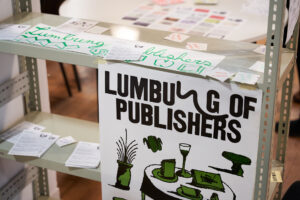 A steel shelf on which many flyers are displayed and a poster with green drawings of a table with a glass and some notebooks.