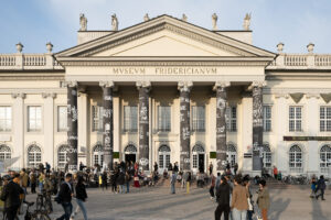 The photo shows the white neoclassical facade of the Fridericianum, with many visitors in front of it. The front of the building is characterized by a portico supported by six Ionic columns which were painted in black with sketches on it.